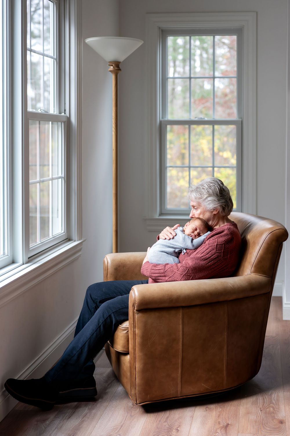 Elderly grandparent sitting in leather armchair holding sleeping newborn baby near bright windows.