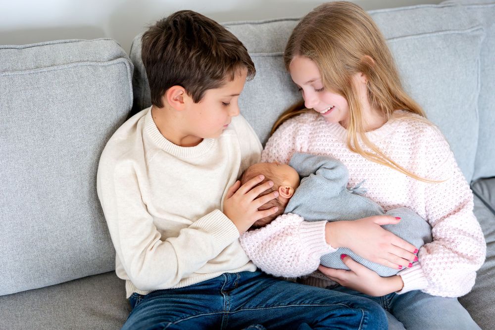Two young siblings sitting on a gray sofa together holding a sleeping newborn baby.