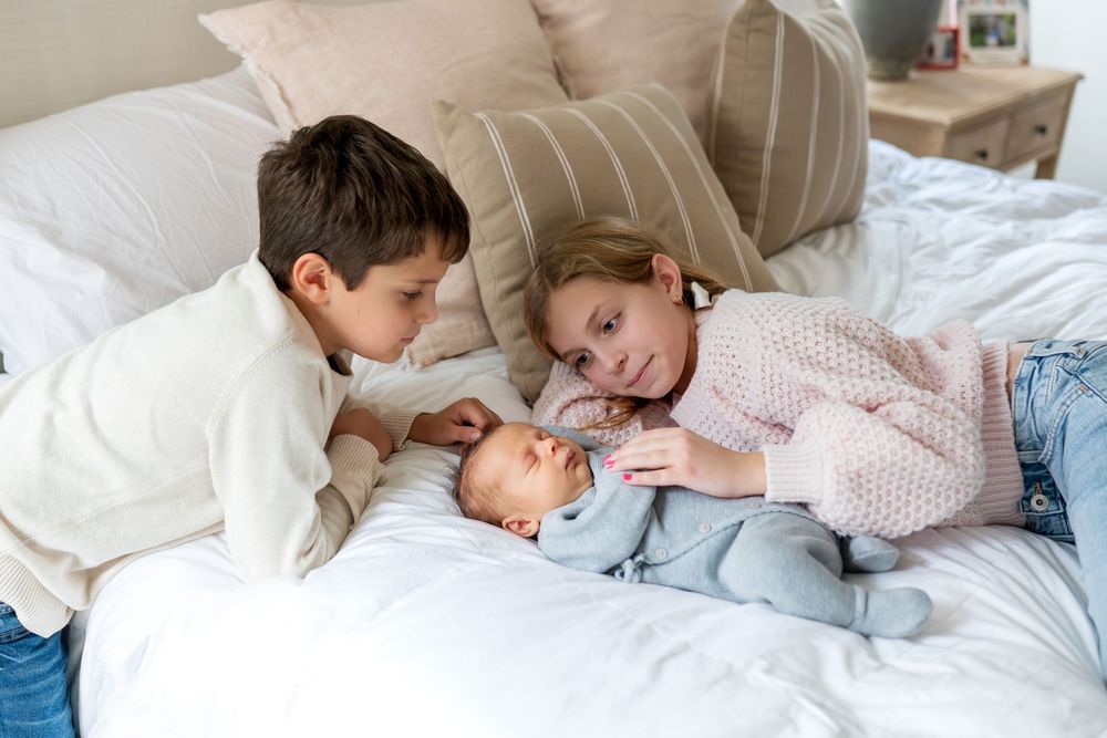 Two young siblings on white bed curiously looking at their sleeping newborn baby.