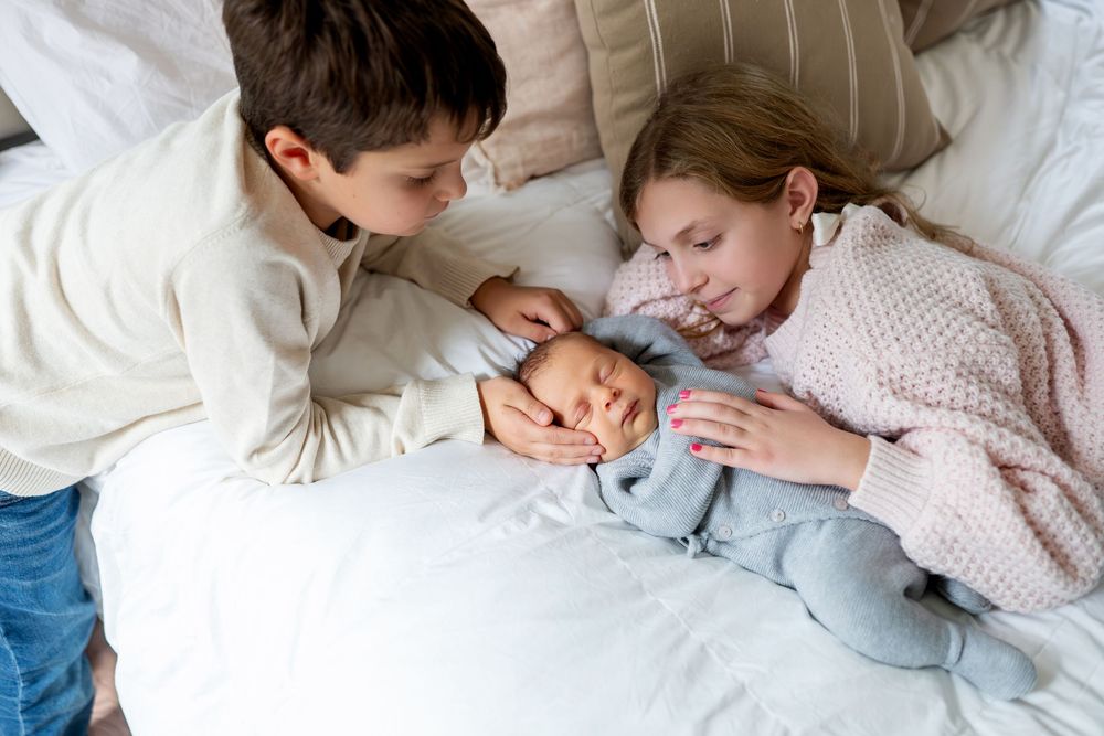 Two children in white and pink sweaters cuddle with newborn baby on white bed.