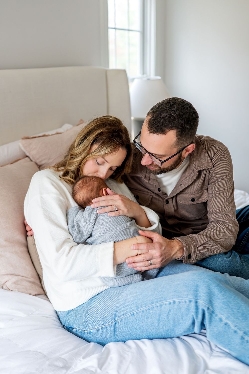 New parents sit closely together on white bed lovingly holding their sleeping newborn.