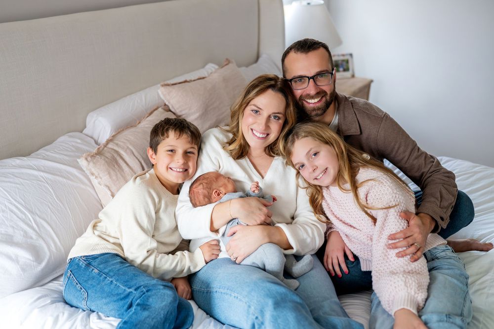 Happy family of five posing on a bed, parents with two kids and a newborn baby smiling at the camera.