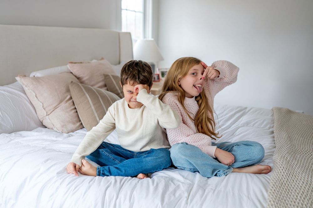 Two young siblings sitting cross-legged on a white bed, laughing and playing together in casual clothes.