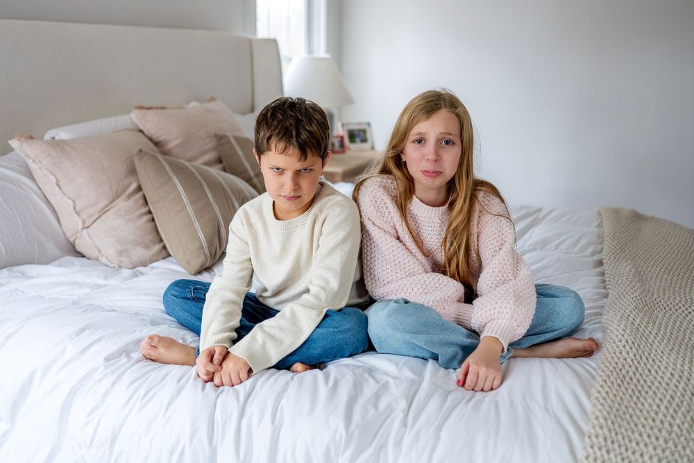 Two children sitting together on a white bed looking sad and upset, boy and girl in casual clothing.