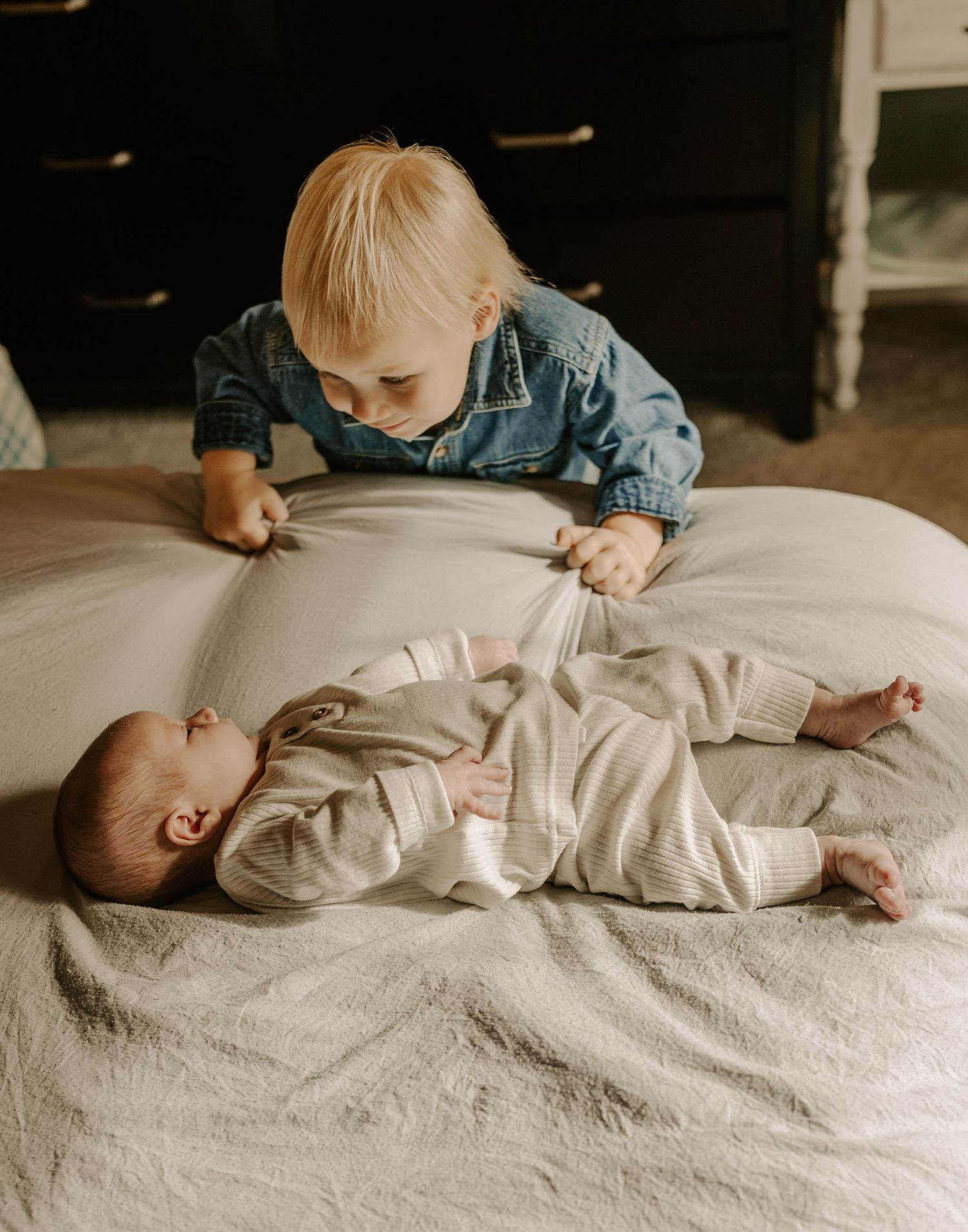 Toddler looking at baby sibling lying on white bedding during photo session.