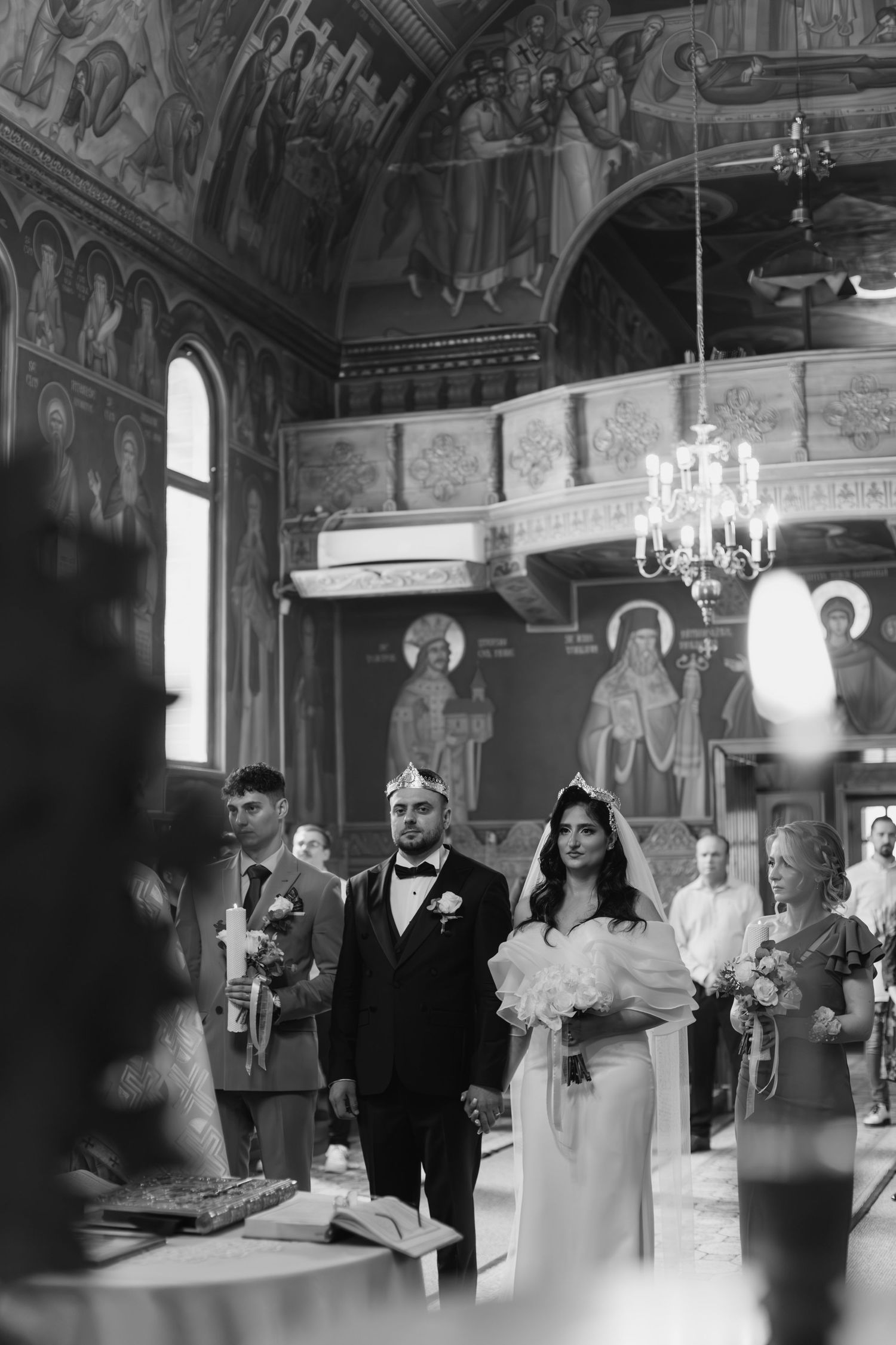 A traditional wedding ceremony takes place inside an ornate Orthodox church with religious murals and a crystal chandelier.