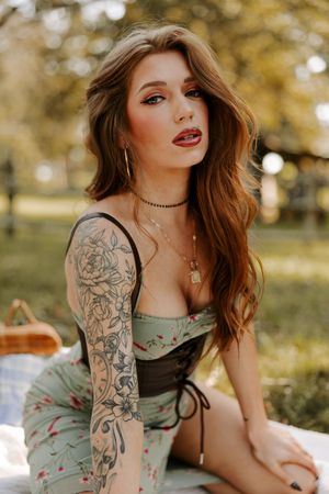 Woman in vintage-style dress with floral pattern and sleeve tattoos poses on white blanket in sunlit outdoor setting.