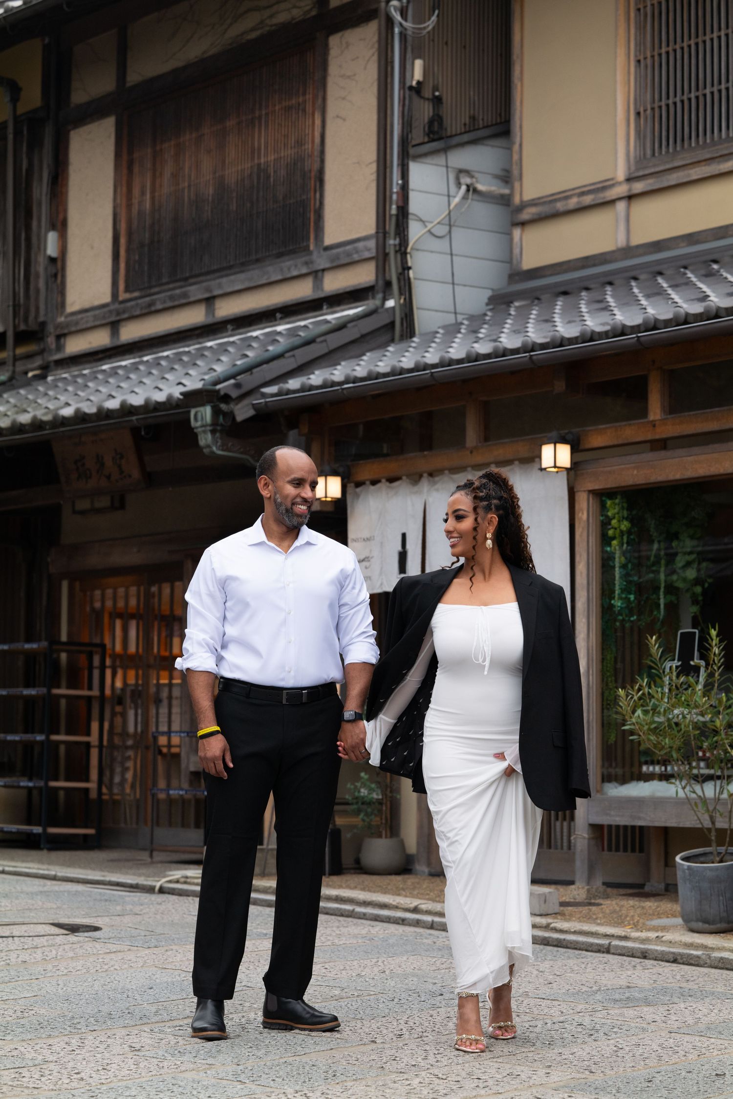 Couple strolling past historic Japanese wooden buildings, man in white shirt, woman in white dress with black blazer.