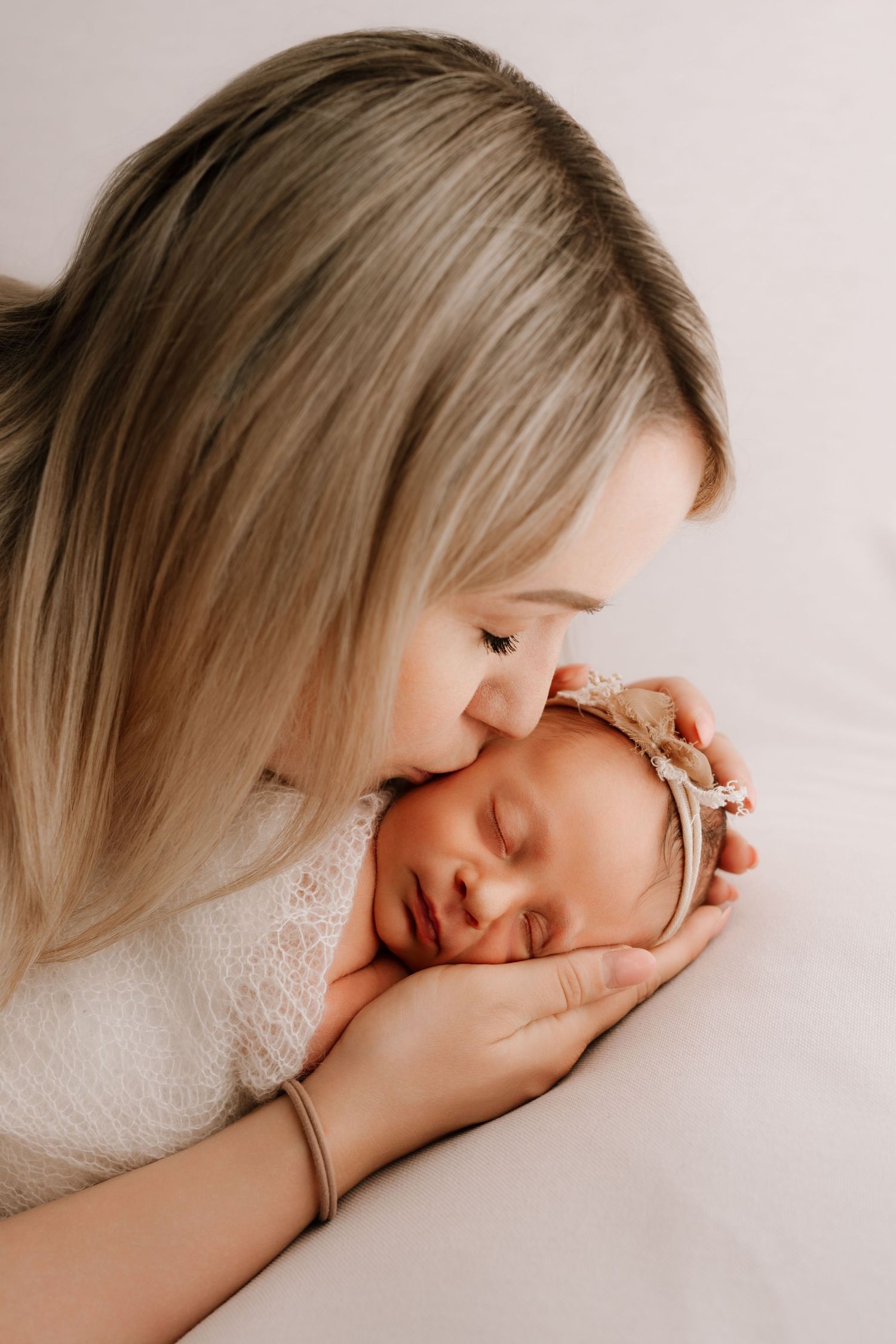 A mother tenderly kisses her sleeping newborn baby girl wearing a delicate headband.