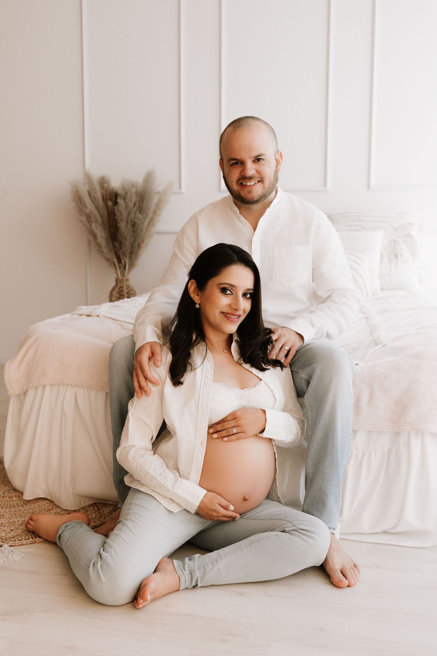 A smiling couple poses together during a maternity photoshoot, with the pregnant woman sitting cross-legged on the floor.