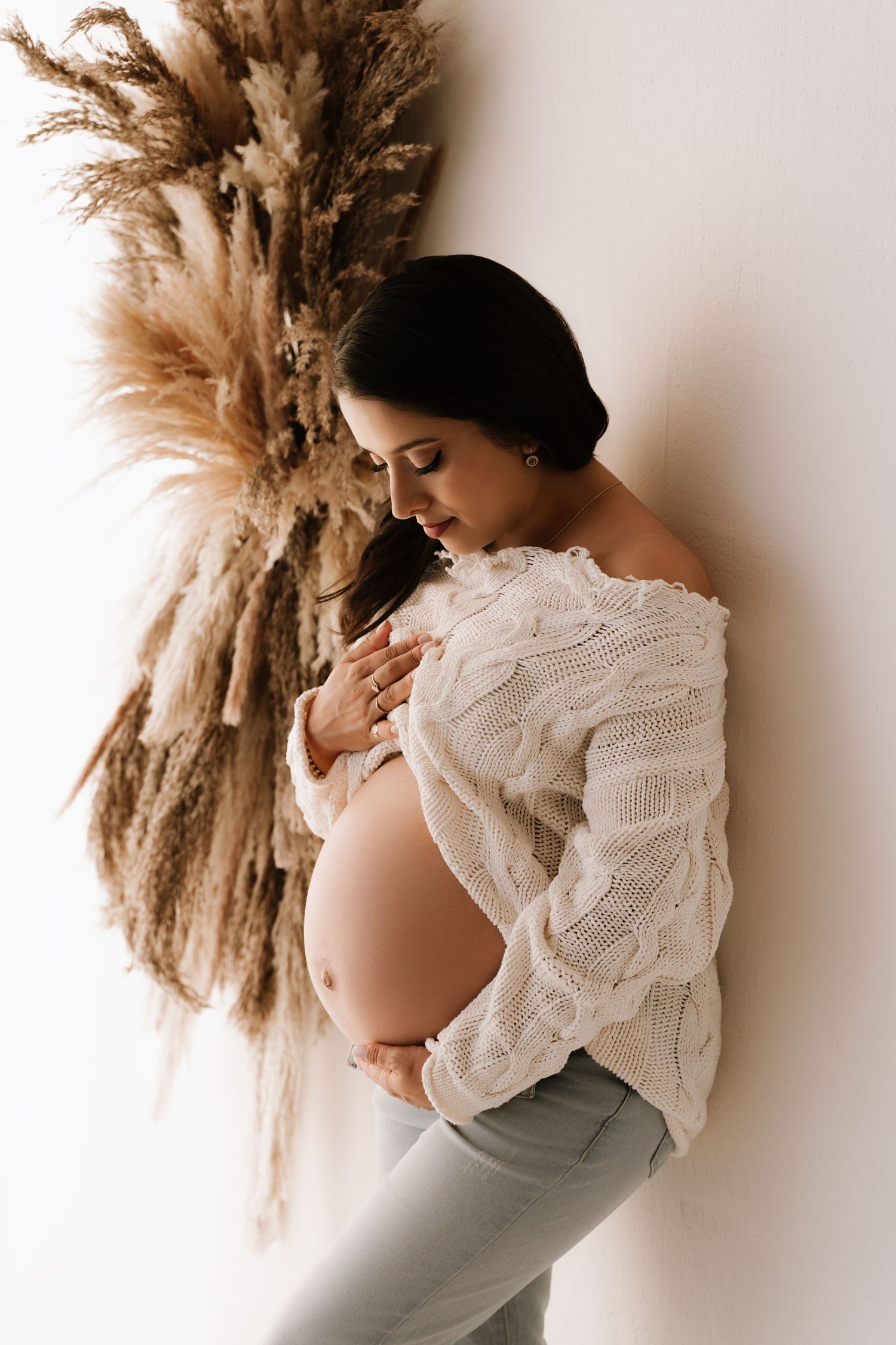 A pregnant woman in a white knit sweater cradles her baby bump beside dried pampas grass decor.