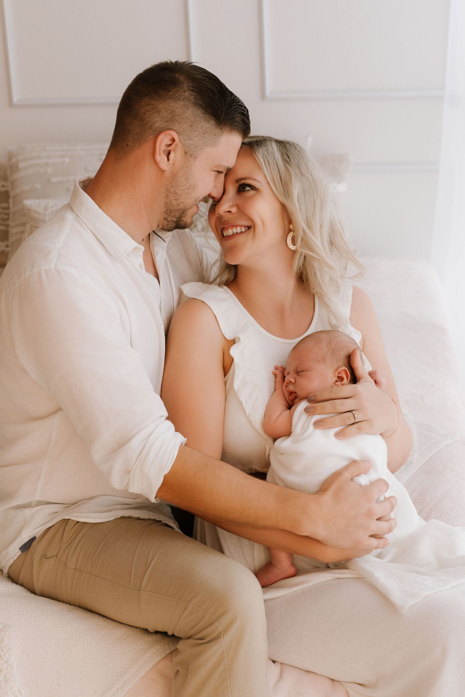 A happy couple in white outfits sits on a bed, lovingly holding their newborn baby.