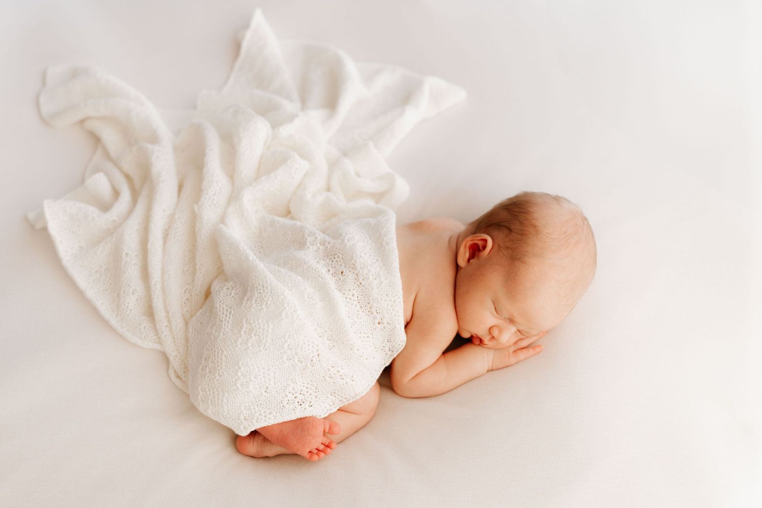 Newborn baby sleeping curled up under a soft white knit blanket on white surface.