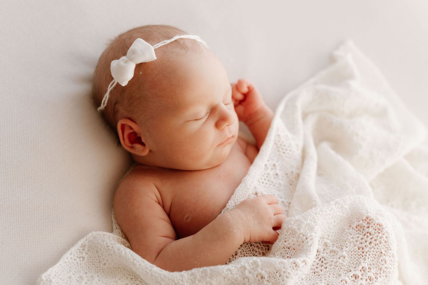 Newborn baby girl with white bow headband sleeping under a white lace blanket.