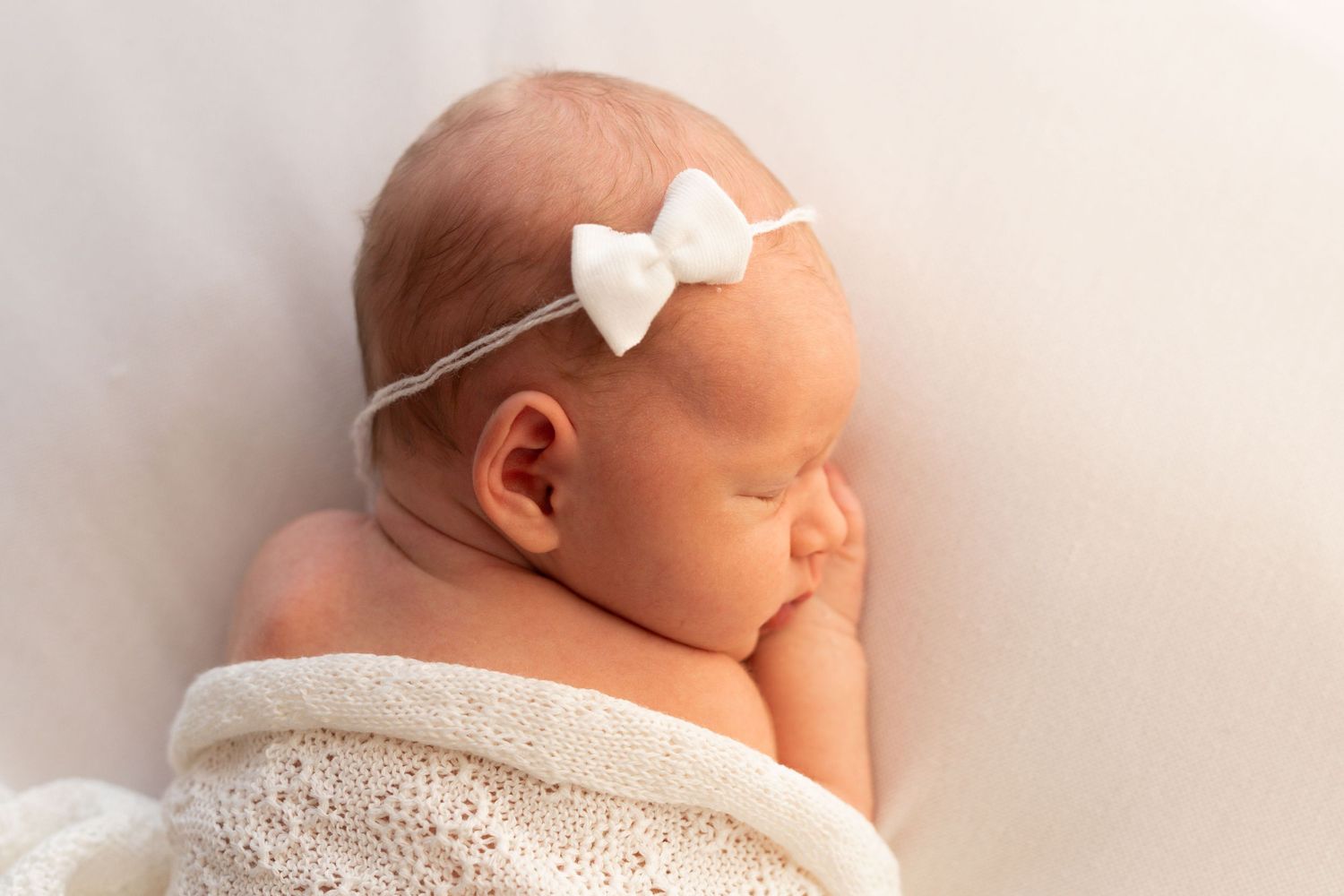 Newborn baby girl with white bow headband sleeping peacefully wrapped in cream knit.