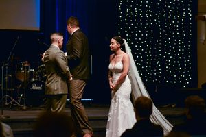 A wedding ceremony takes place on stage with a backdrop of twinkling string lights and musical instruments.