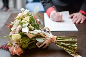 A delicate bridal bouquet with roses and greenery tied with ribbon, with document signing in background.
