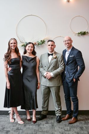 Wedding party poses together against neutral wall wearing formal black dresses and suits.