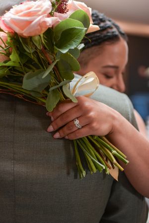 Pink roses and greenery are held in a bouquet with an engagement ring visible on the hand.