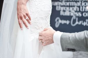 Close-up detail of lace wedding dress with hands showing rings against a chalkboard wall.