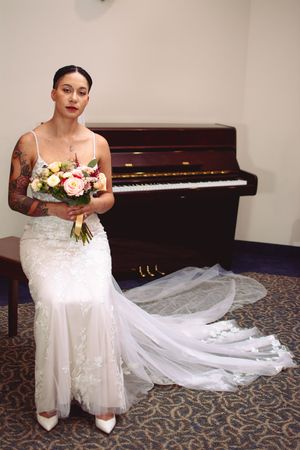 A bride in a white wedding dress with a long train stands beside a piano holding a floral bouquet.