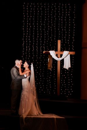 Wedding couple embraces in front of illuminated cross and string lights.