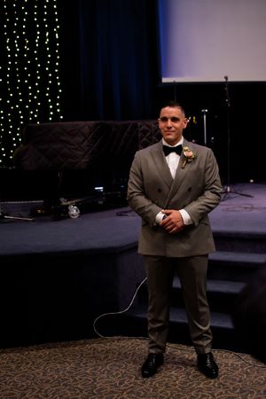 A person in a gray suit stands on a stage with twinkling string lights in the background.