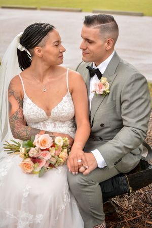 Newlyweds share intimate moment on rustic bench with peach bouquet.