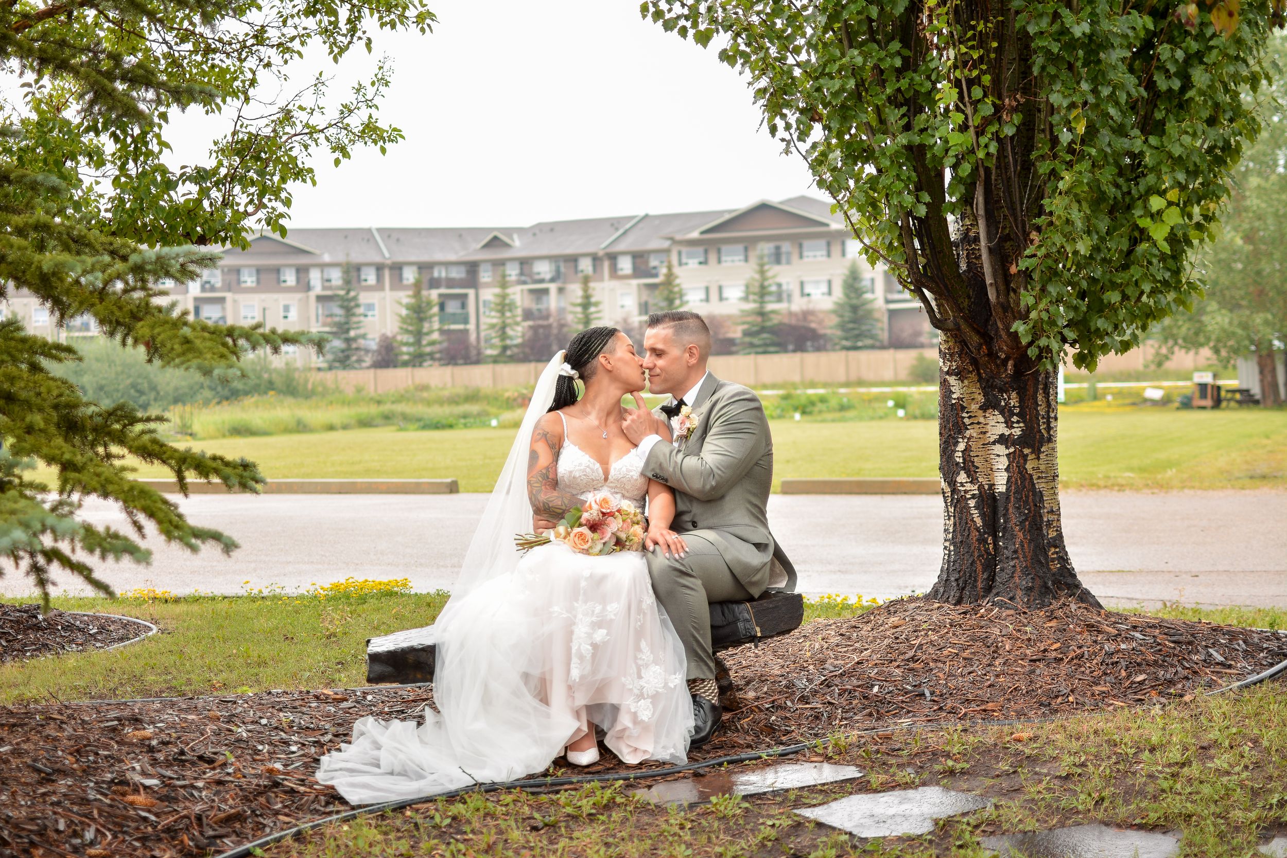 Outdoor wedding photography session with couple posing under green trees near a building.