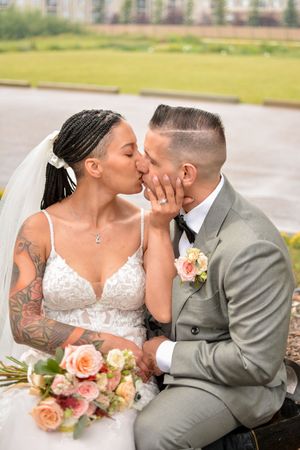 Newlywed couple in grey suit and white dress embrace while holding coral-colored bouquet outdoors.
