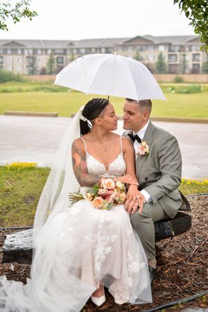 A romantic wedding couple shares an intimate moment under a white umbrella with apartment buildings in the background.