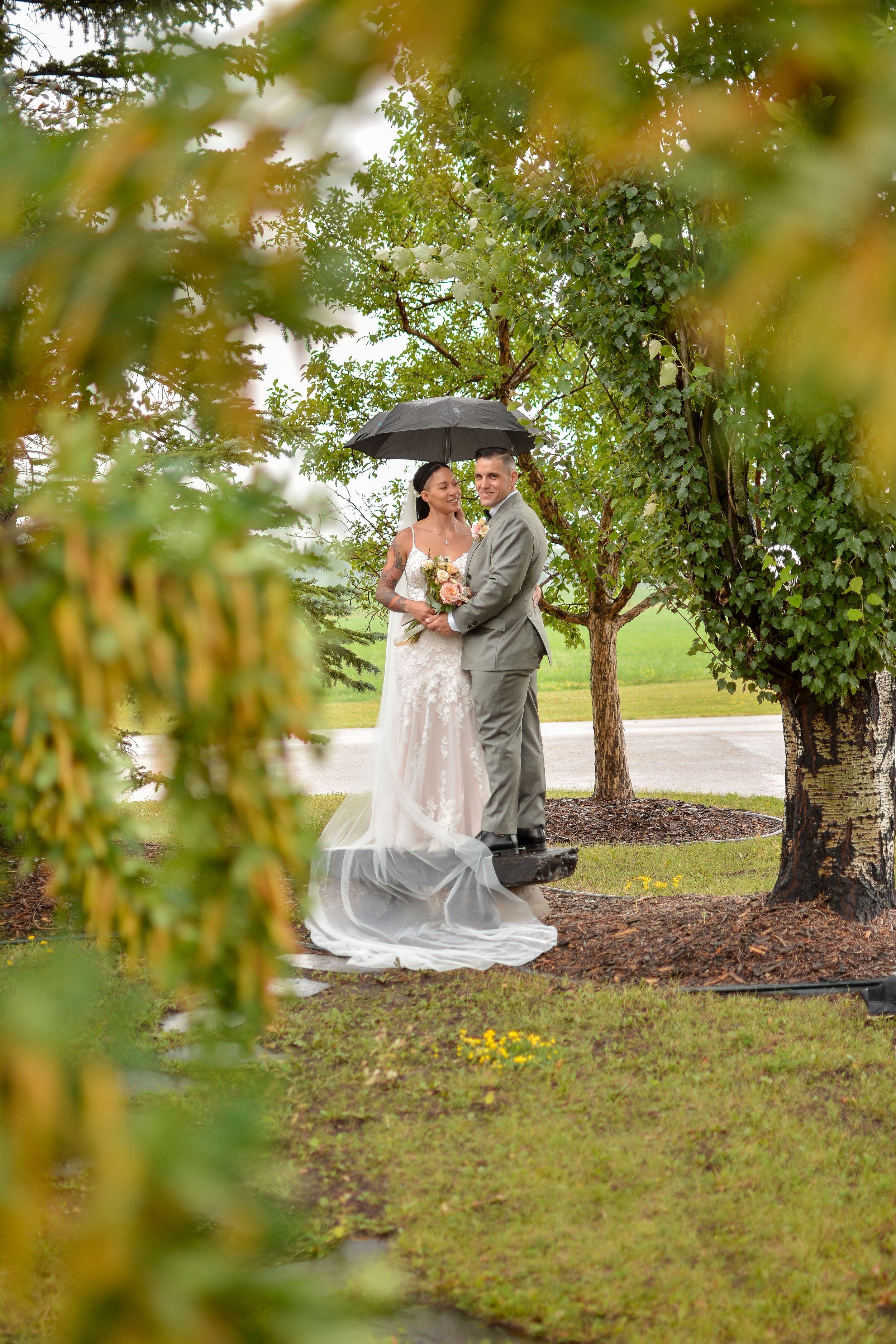 Wedding couple shares a tender moment under green trees in an outdoor garden setting.