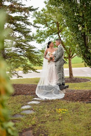 A wedding couple poses together on a grassy hillside near trees in a romantic outdoor setting.