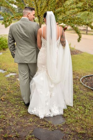 Wedding couple walking together on garden path with flowing white veil trailing behind.