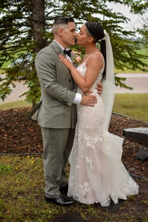 Romantic wedding couple embraces under evergreen trees in outdoor setting.