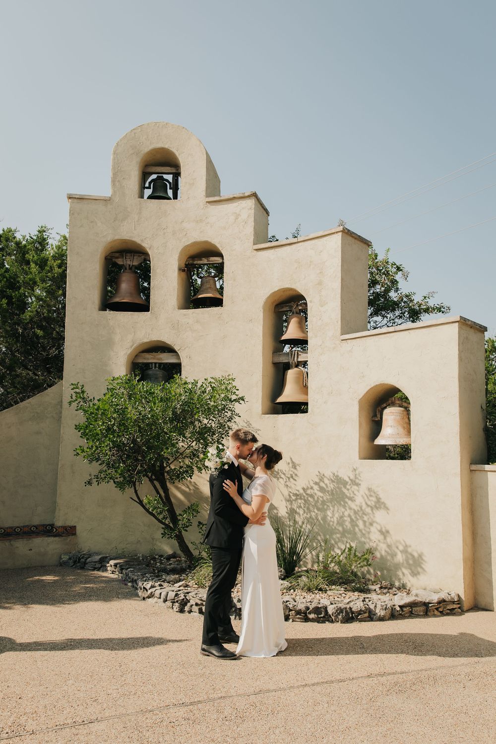 A couple stands together against a southwestern style bell tower with multiple arches and bells in the desert landscape.