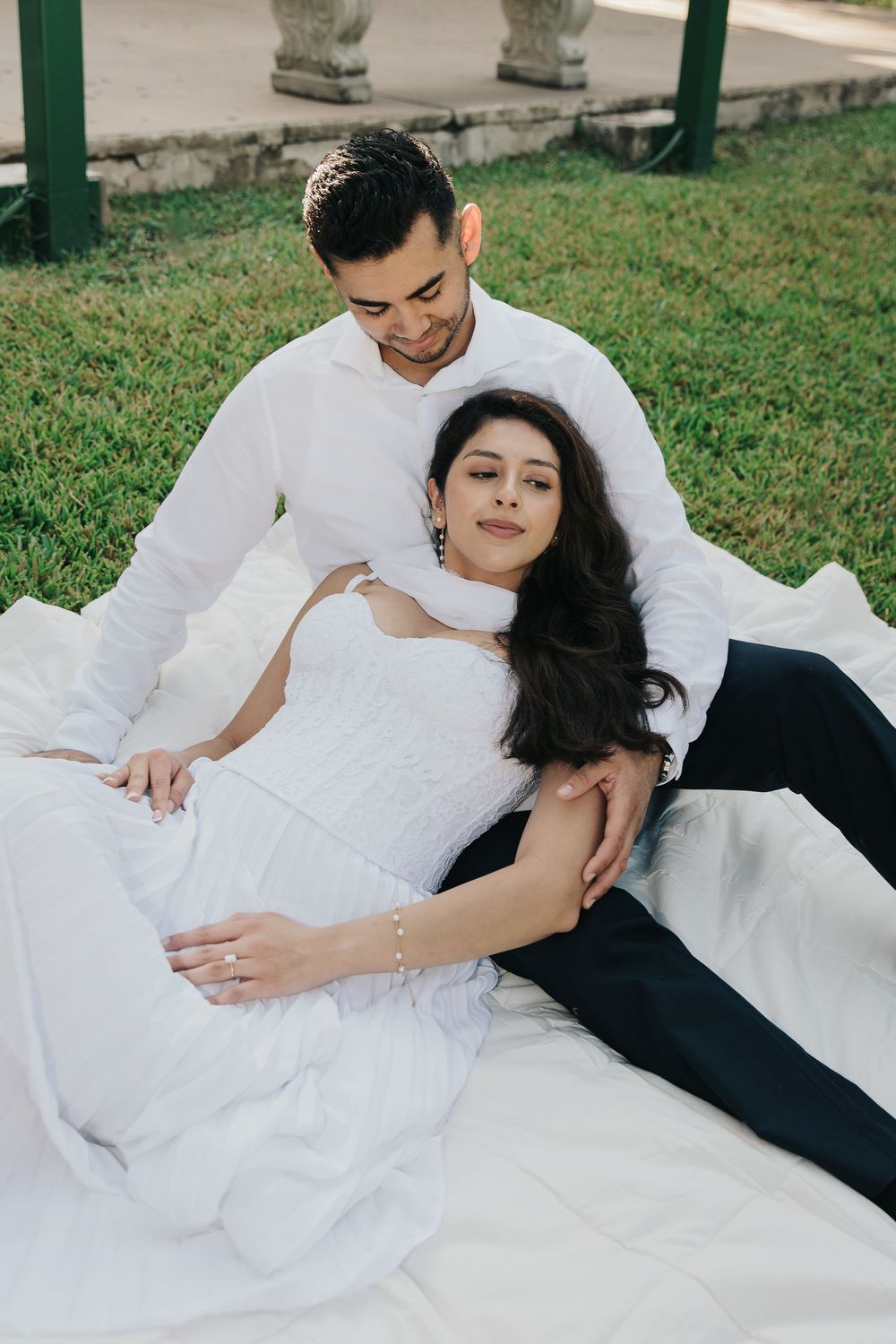 A bride and groom in white and black attire share intimate moments while laying on grass outdoors.