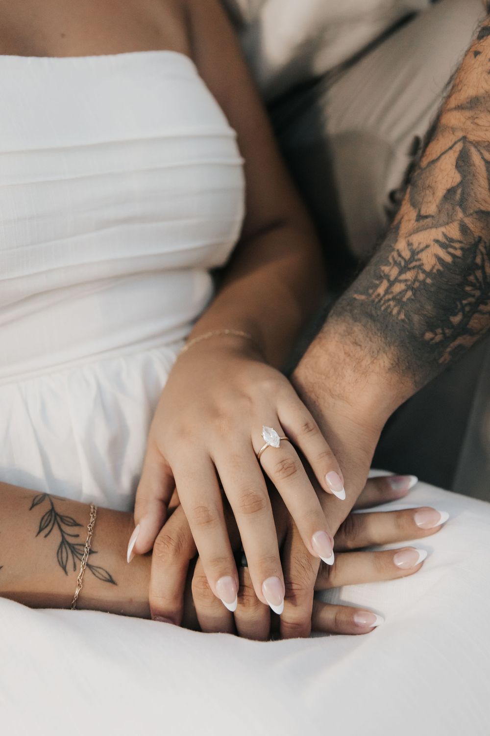 Close-up of hands with delicate tattoos and engagement ring against white fabric showing romantic wedding moment.