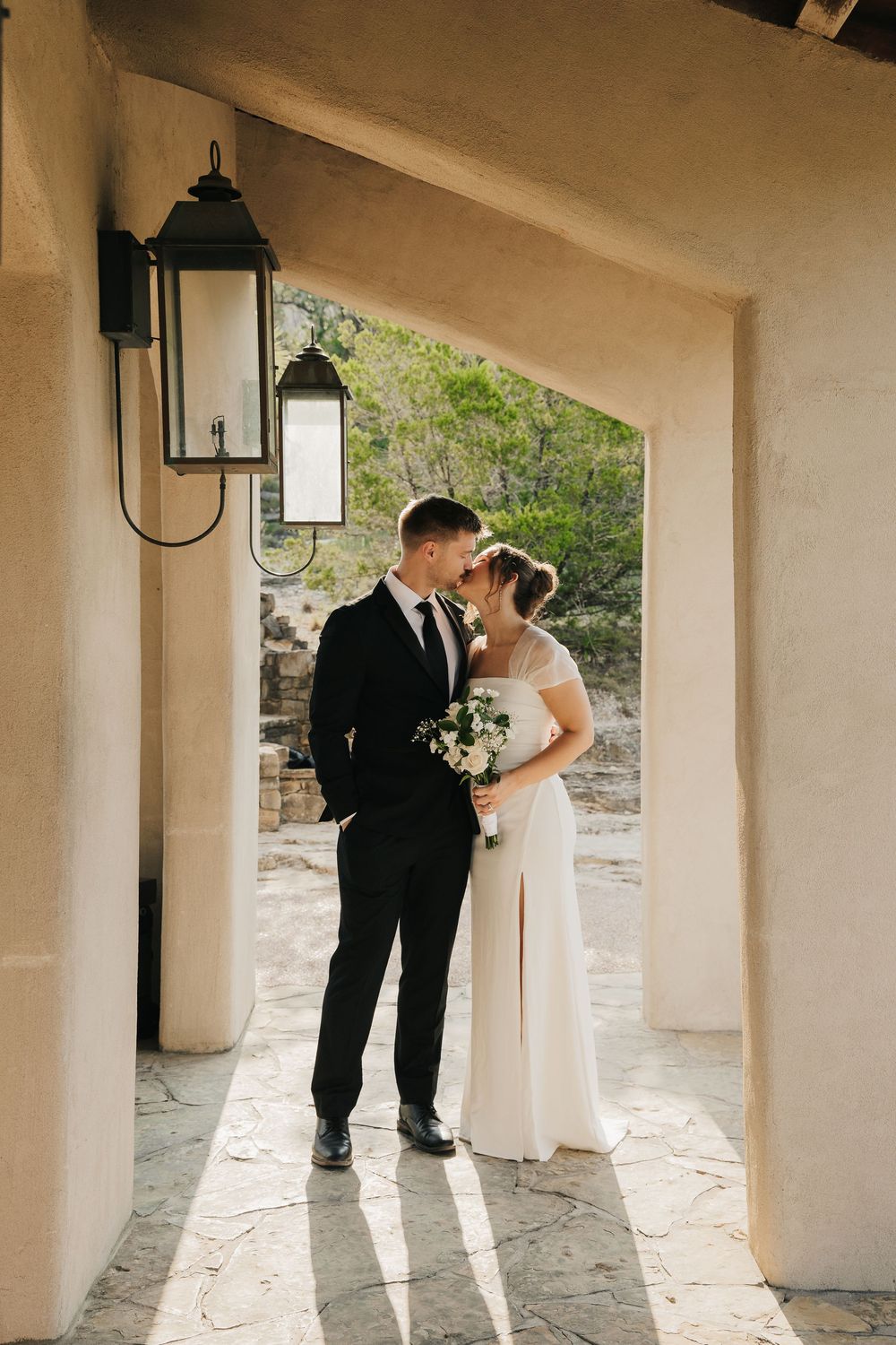 A couple in wedding attire stands beneath a colonnade of cream-colored pillars, creating a romantic architectural frame.