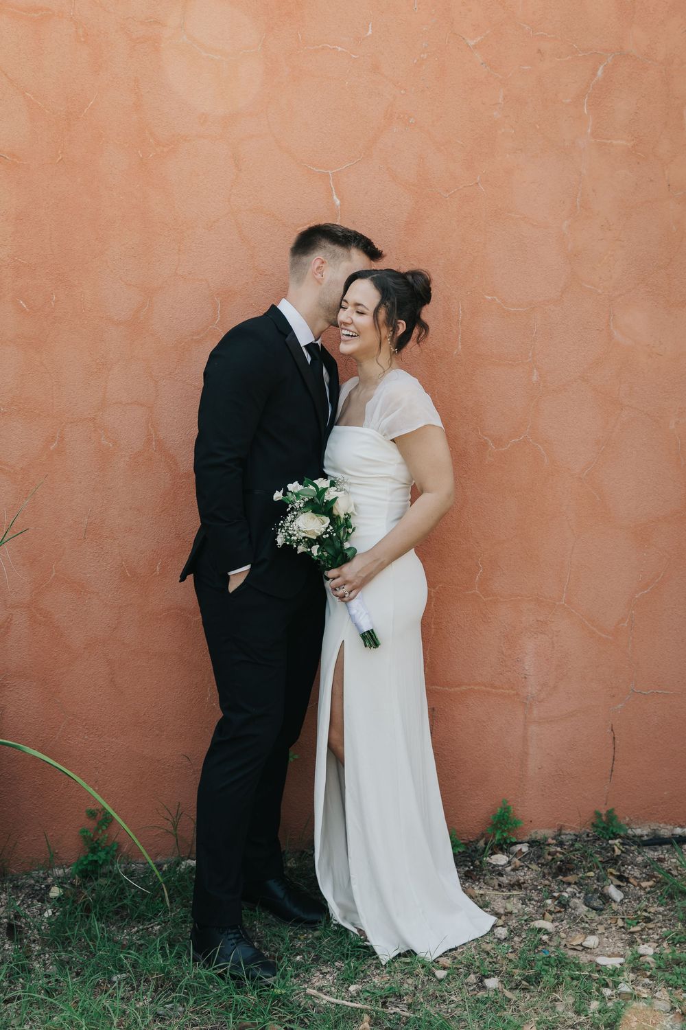 A couple in wedding attire share an intimate moment against a terracotta colored wall outdoors.