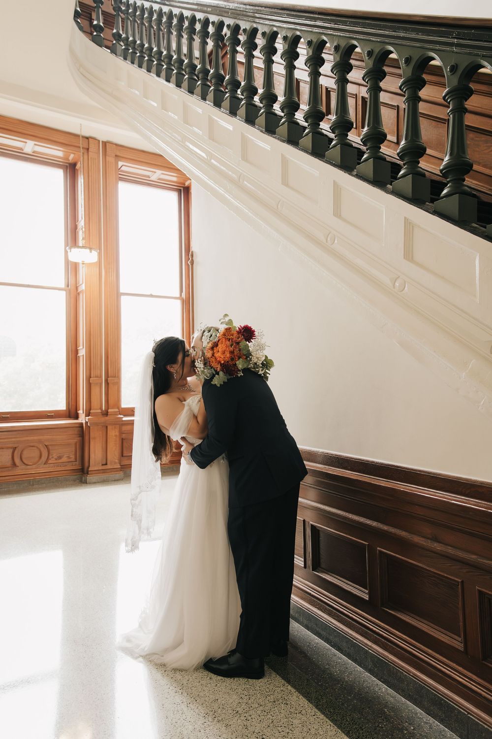 Wedding couple embracing on a wooden staircase with ornate black railings and natural window light.