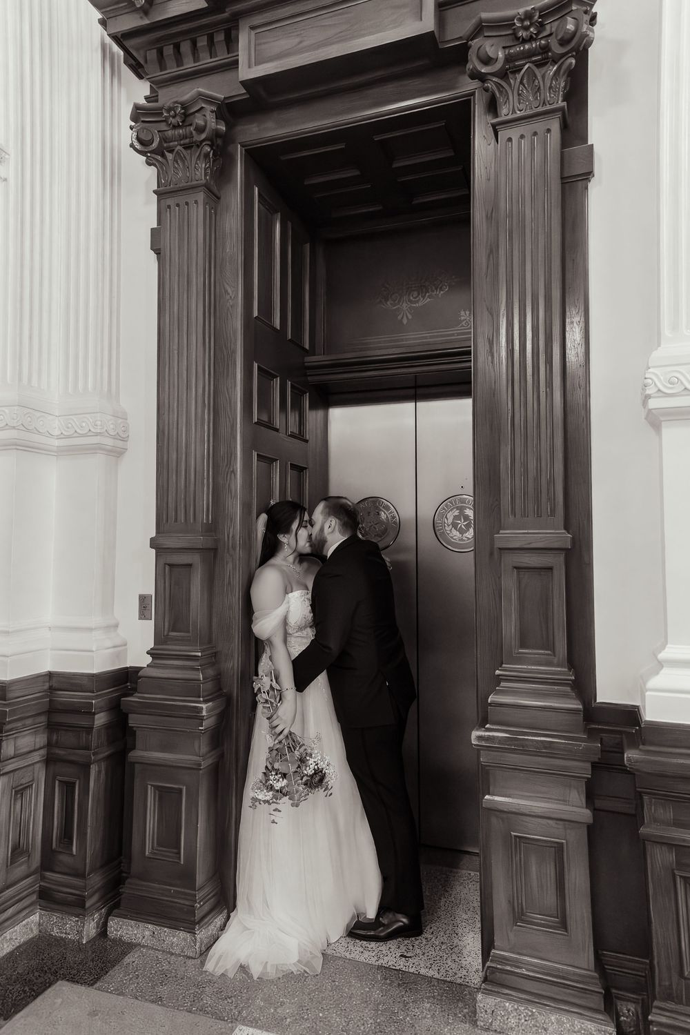 A couple shares an intimate moment in an elegant doorway with ornate wooden columns and architectural details.