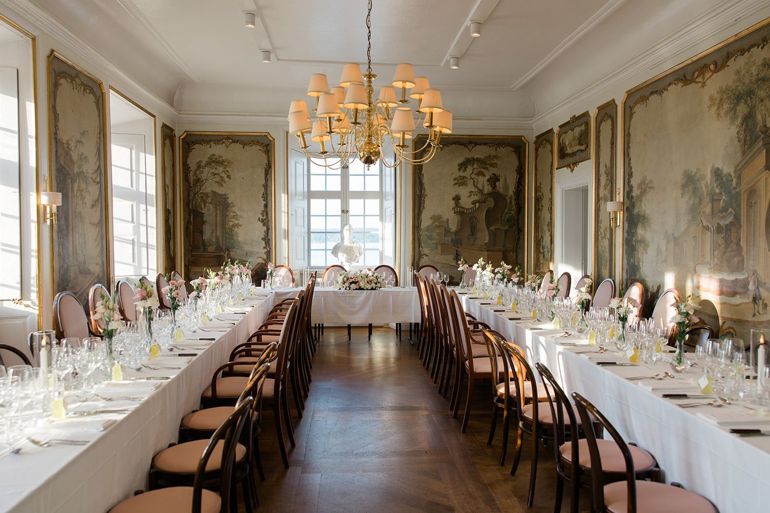 Elegant banquet hall with long dining tables, ornate chandeliers, and classic white decor in a historic setting.