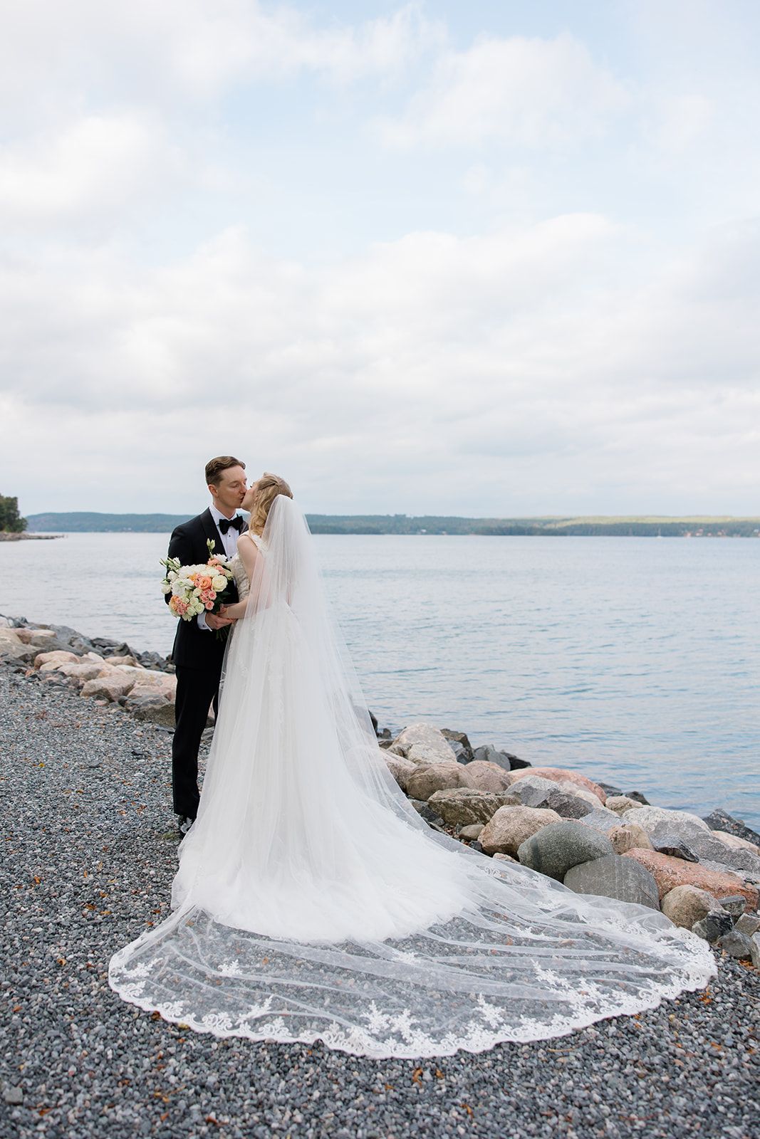 Newlywed couple embracing on rocky beach shore with flowing white wedding dress train and ocean view in background.