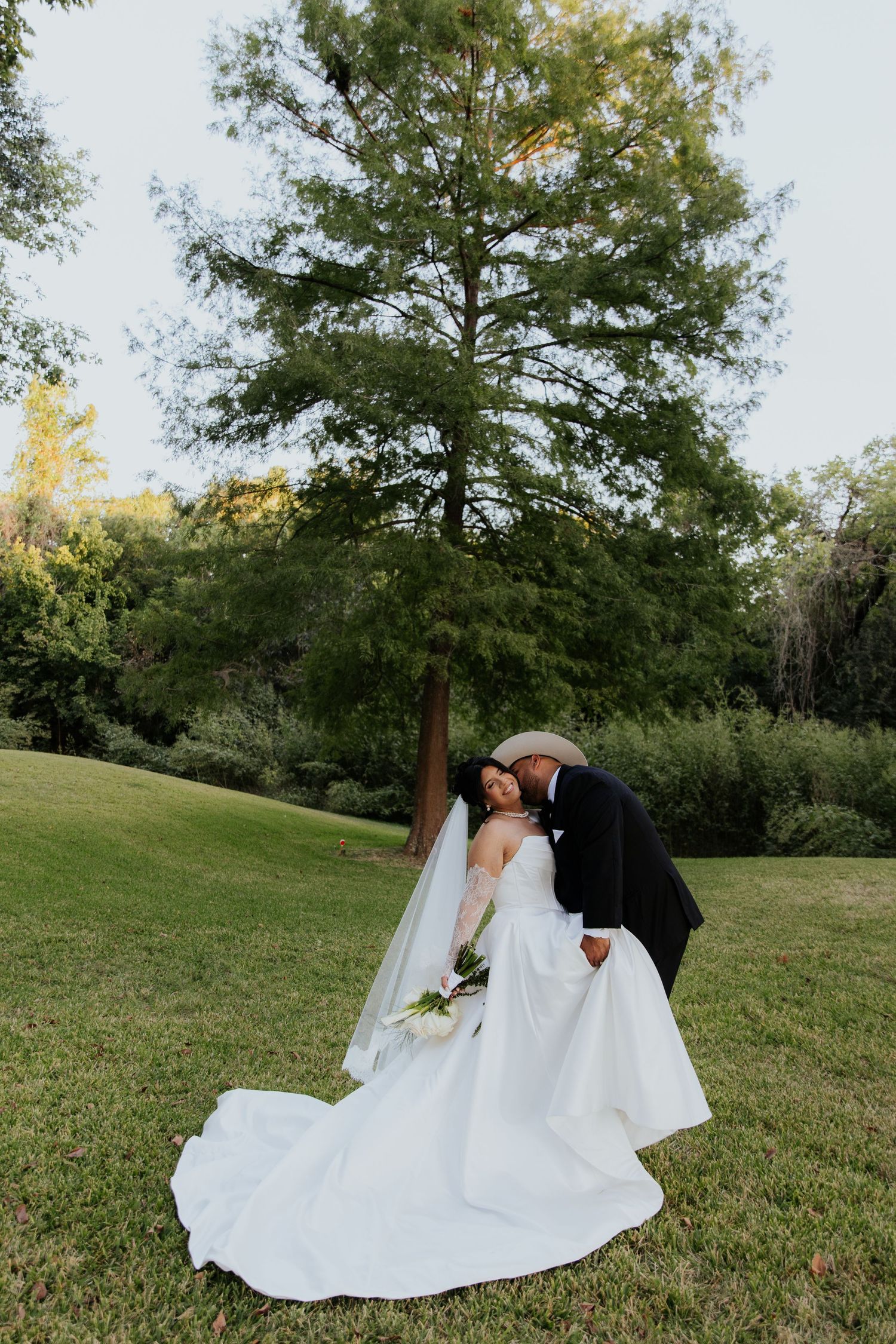 Wedding couple shares intimate moment on grassy hillside under large tree.