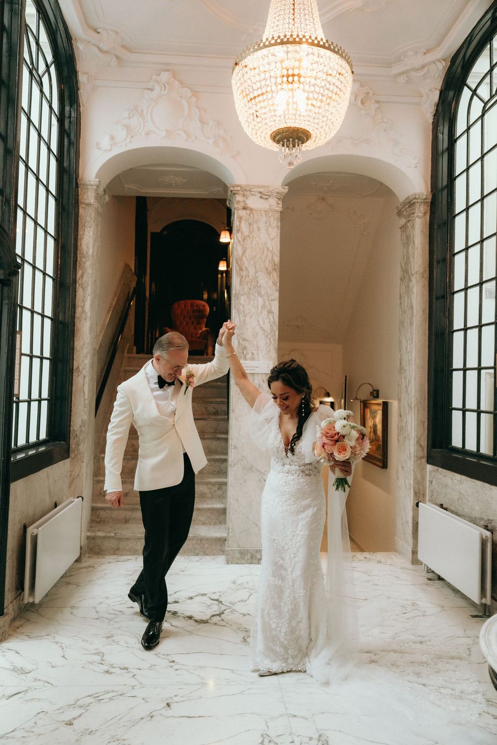 Elegant wedding couple poses in a luxurious marble hallway with crystal chandeliers and ornate columns.