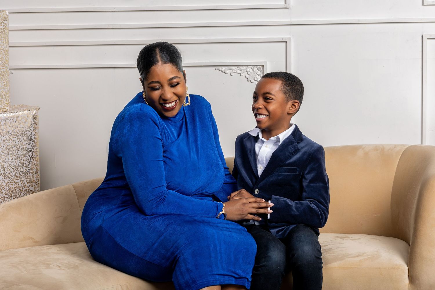 Woman in blue outfit laughing with young boy in velvet blazer on a couch.