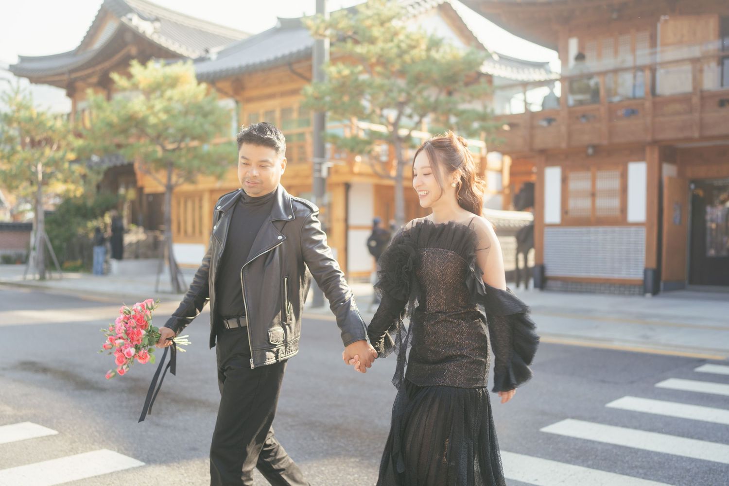 A couple in dark clothing walking together through a traditional Japanese temple complex on a sunny day.
