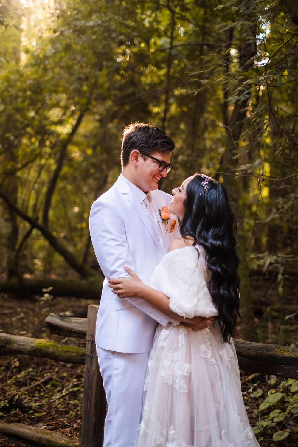 A bride and groom in white attire share intimate moments in a sunlit forest setting during their photoshoot. in the redwoods in santa cruz