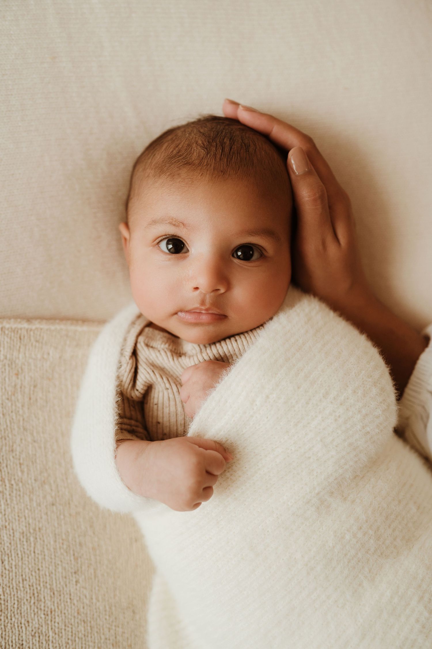 A baby in a cozy cream sweater lies on a light-colored surface while a hand gently rests on their head.